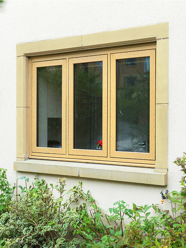 A three-panel flush sash window set into a cream-colored wall, surrounded by a stone border. The right panel is slightly open, and reflections of greenery and a house are visible in the glass. 