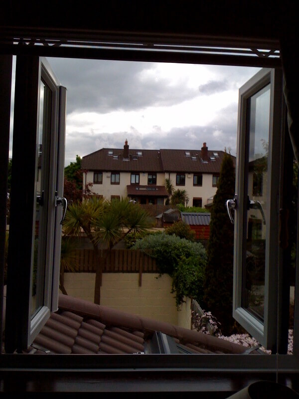 A fully open French window with white frames, showing a view of a suburban neighborhood with houses, palm trees, and a garden. The sky is cloudy, and rooftops and greenery are visible in the background.